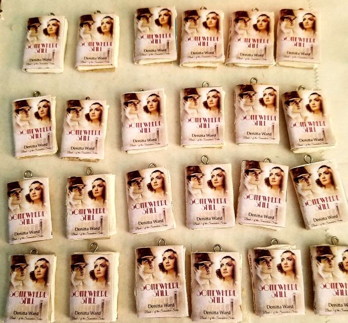 Twenty-four finished Somewhere Still mini-book charms laid out in rows, each with a small metal screw eye at the top